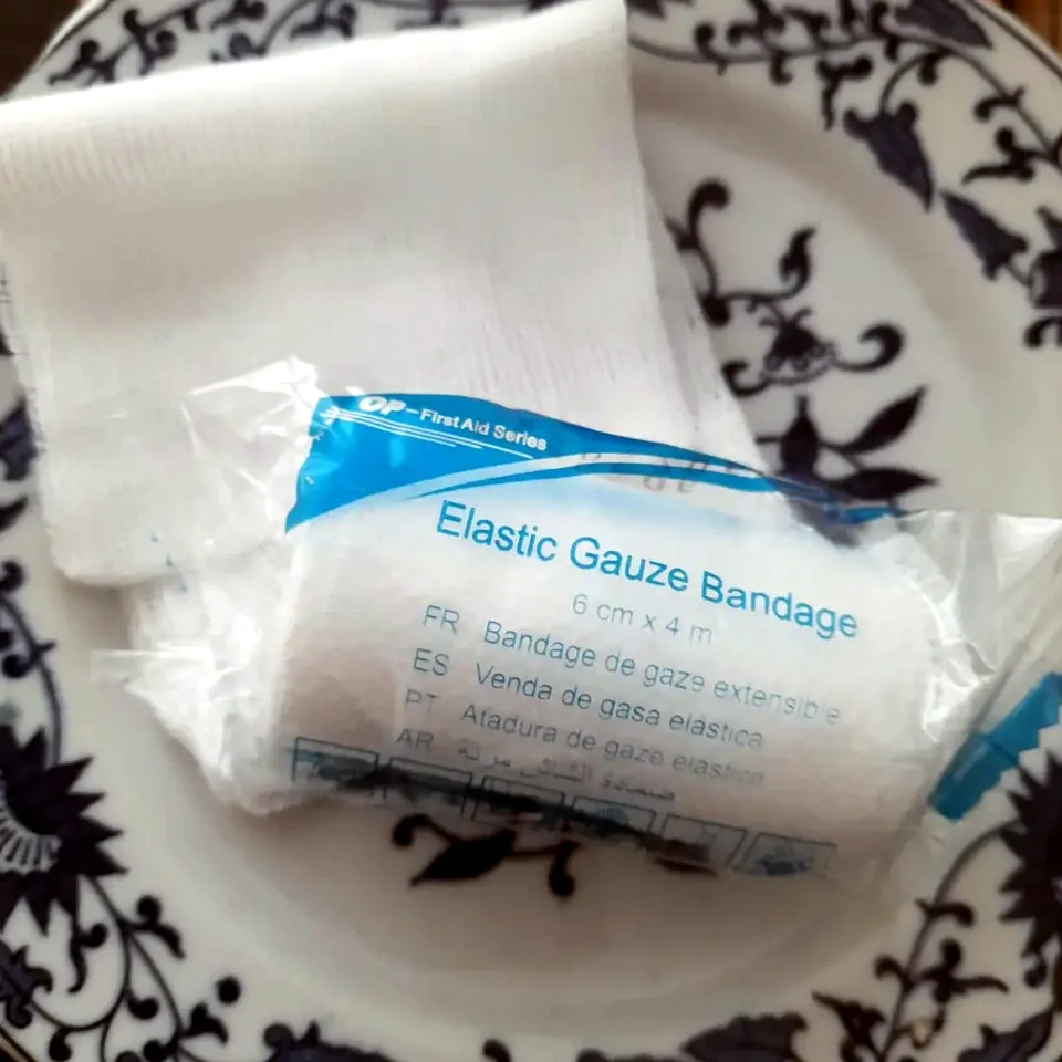 Studio photo: a packet of elastic gauze bandage and a loose piece of gauze on a white plate with a dark floral pattern.