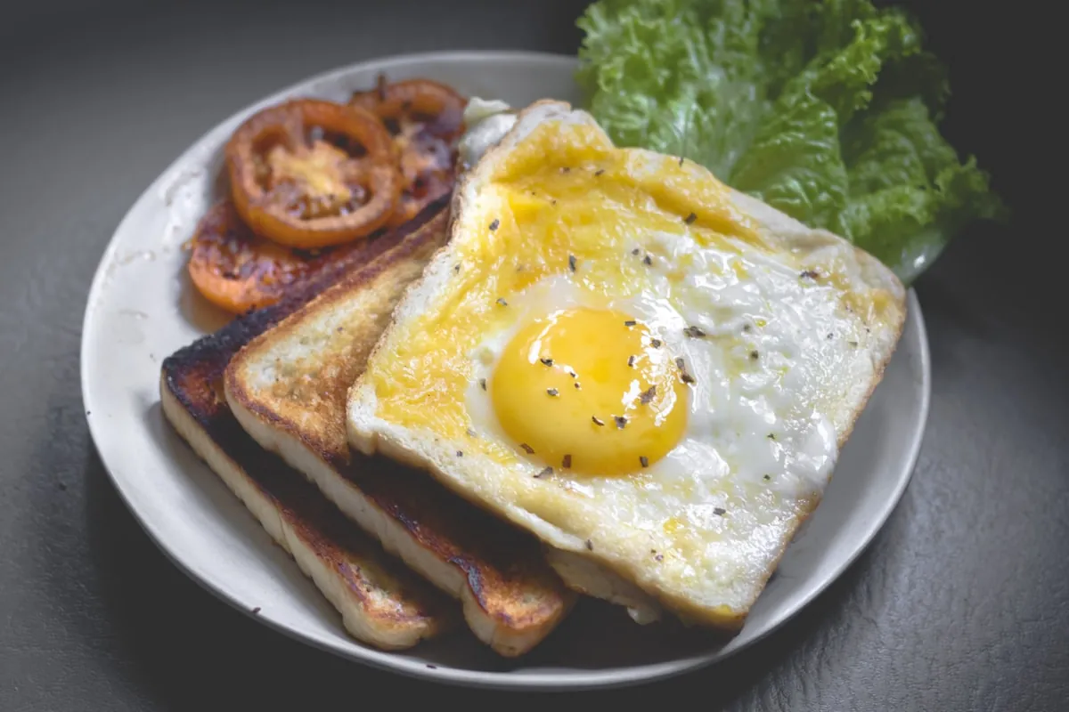 Weißer Teller mit einem Spiegelei auf getoastetem Brot, daneben gegrillte Paradeiserscheiben und Blattsalat.