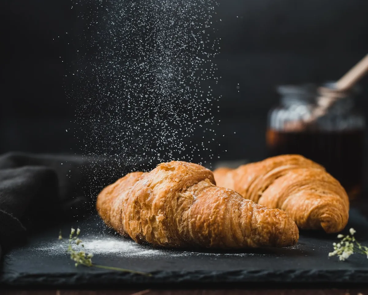 Zwei Butterkipferl auf einer dunklen Schieferplatte, Staubzucker rieselt darauf, dahinter ein Glas Marmelade.