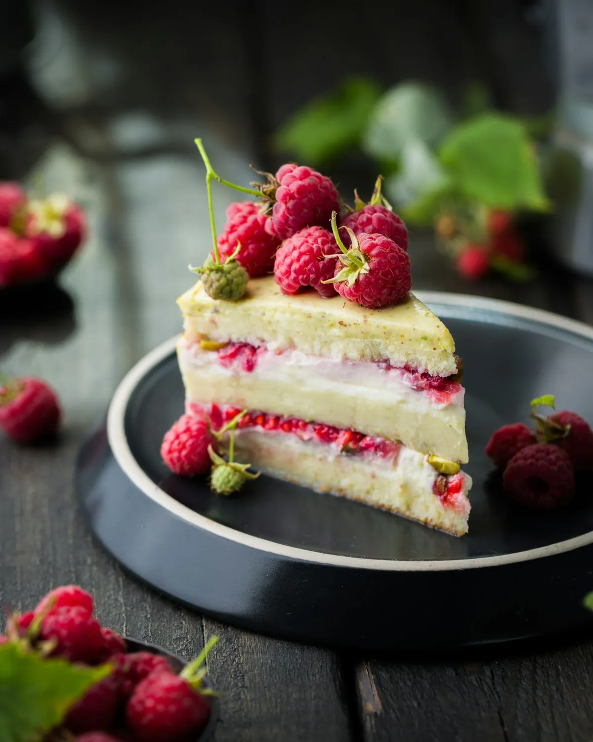 Ein Stück Schichttorte mit heller Creme, Himbeerfüllung und Pistaziendecke, obenauf frische Himbeeren, auf einem dunklen Teller.