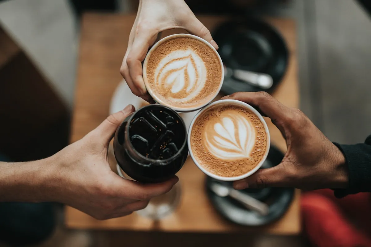 Von oben: drei Hände stoßen über einem Holztisch mit zwei Milchkaffees mit Latte-Art und einem Glas Eiskaffee an.