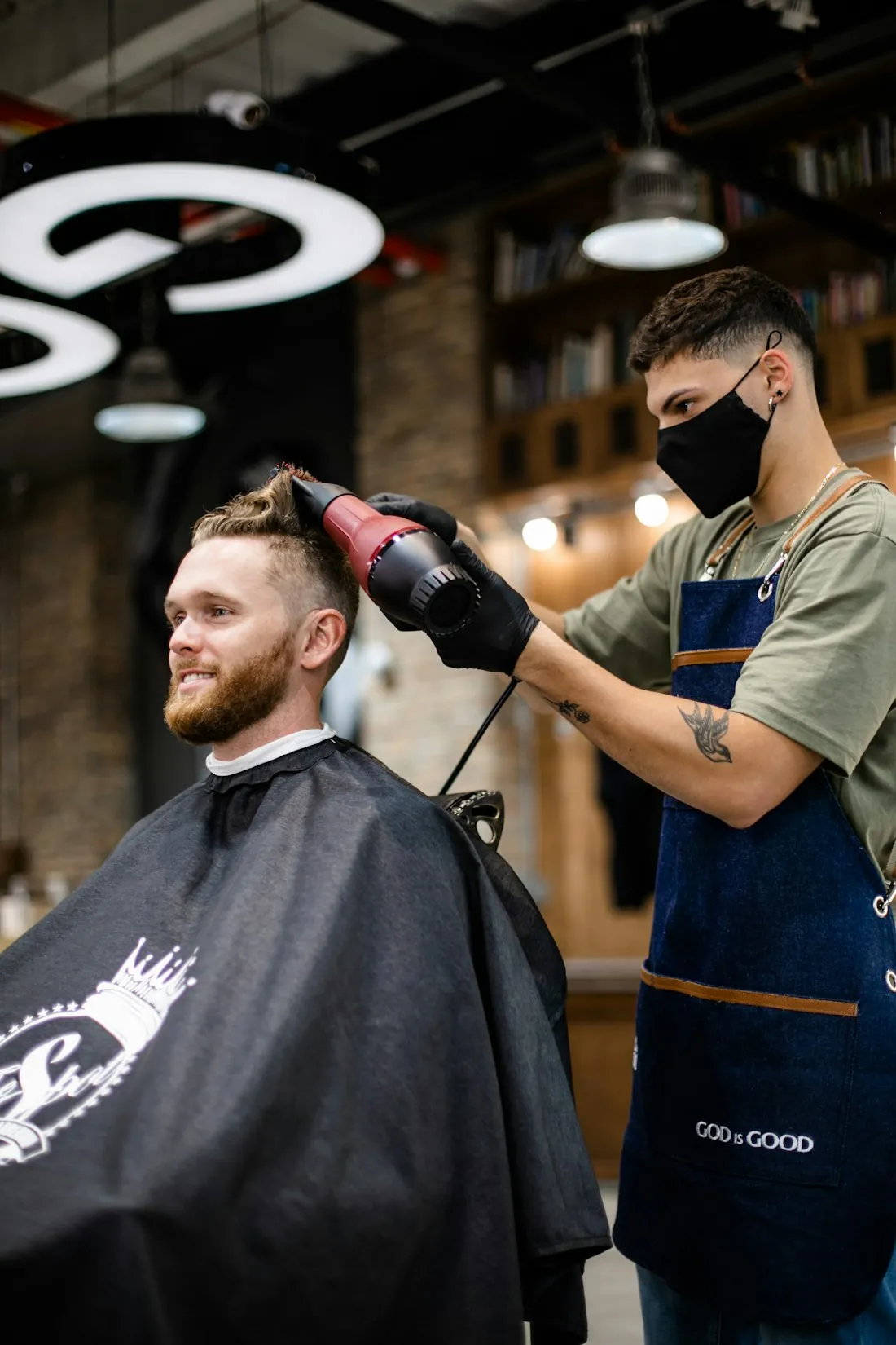Ein Barbier föhnt einem Kunden im Umhang die Haare nach dem Schnitt trocken