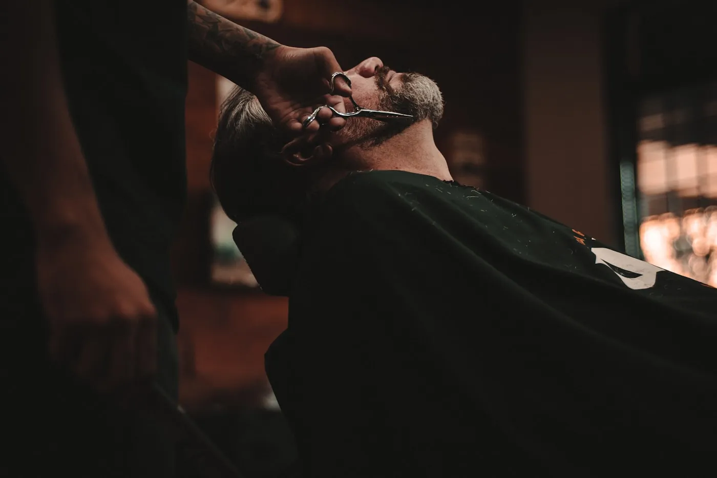 Ein Kunde liegt zurückgelehnt im Barbierstuhl, der Barbier setzt die Schere am Bart an