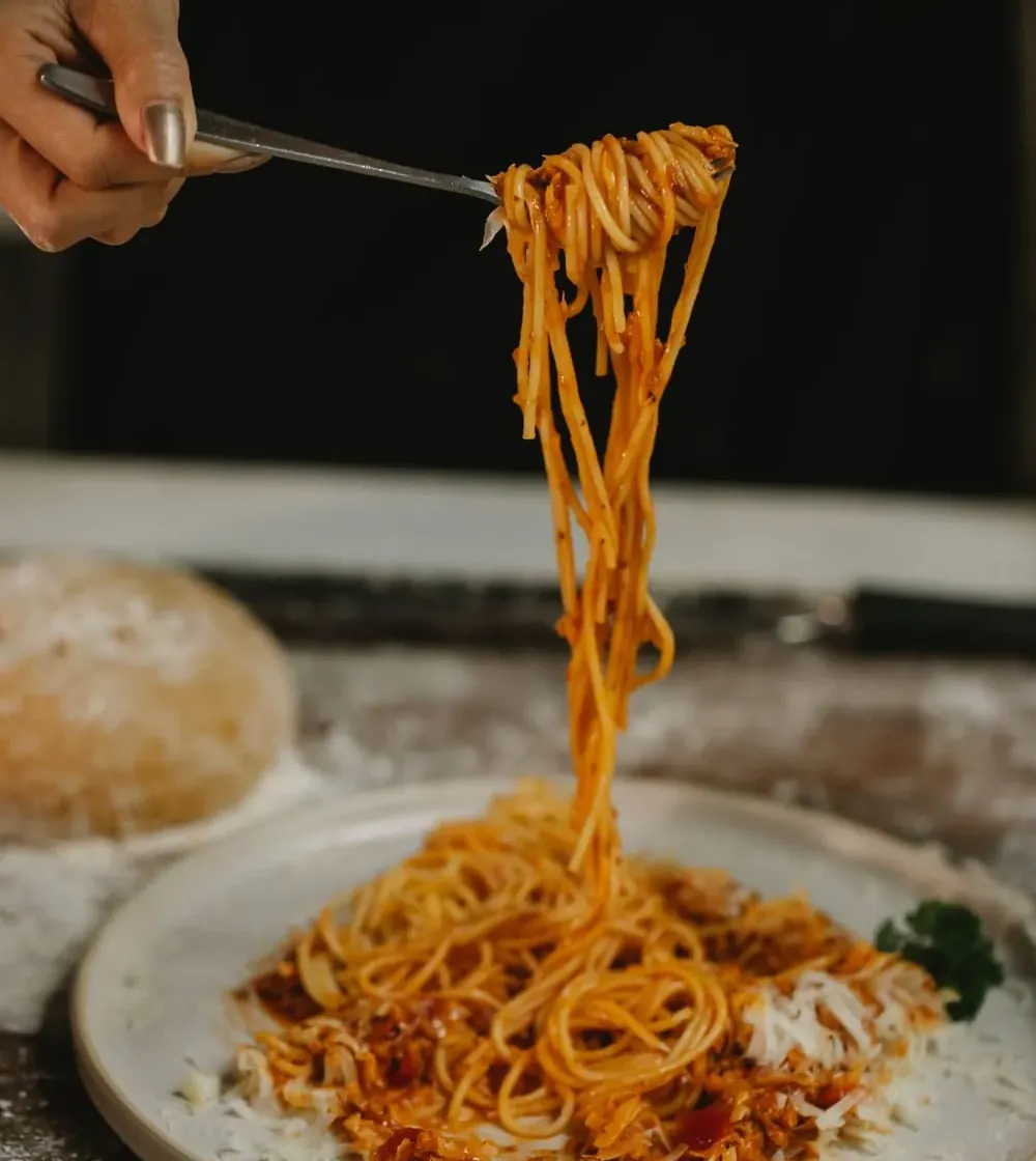 Spaghetti mit Tomatensauce werden auf eine Gabel gedreht, daneben liegt ein Teigballen auf bemehltem Holz
