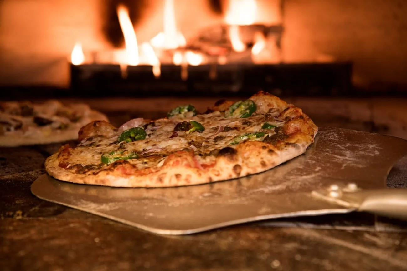 Eine frisch gebackene Pizza mit Peperoni liegt auf dem Metallschieber, dahinter brennt das Feuer im Ofen